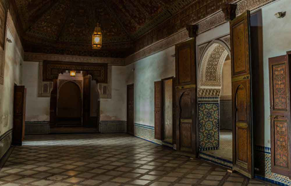 Dar Si Said Museum: A dimly lit room showcasing traditional Moroccan architecture, featuring geometric floor tiles, intricately carved wooden doors, and ornate ceiling patterns.