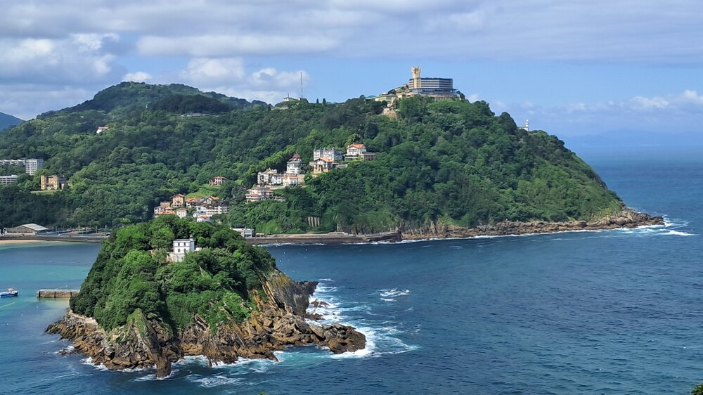 A lush, hilly coastal landscape with scenic buildings nestled into Monte Igueldo overlooking the ocean, representing the kind of natural beauty and vistas that define what San Sebastian is famous for.