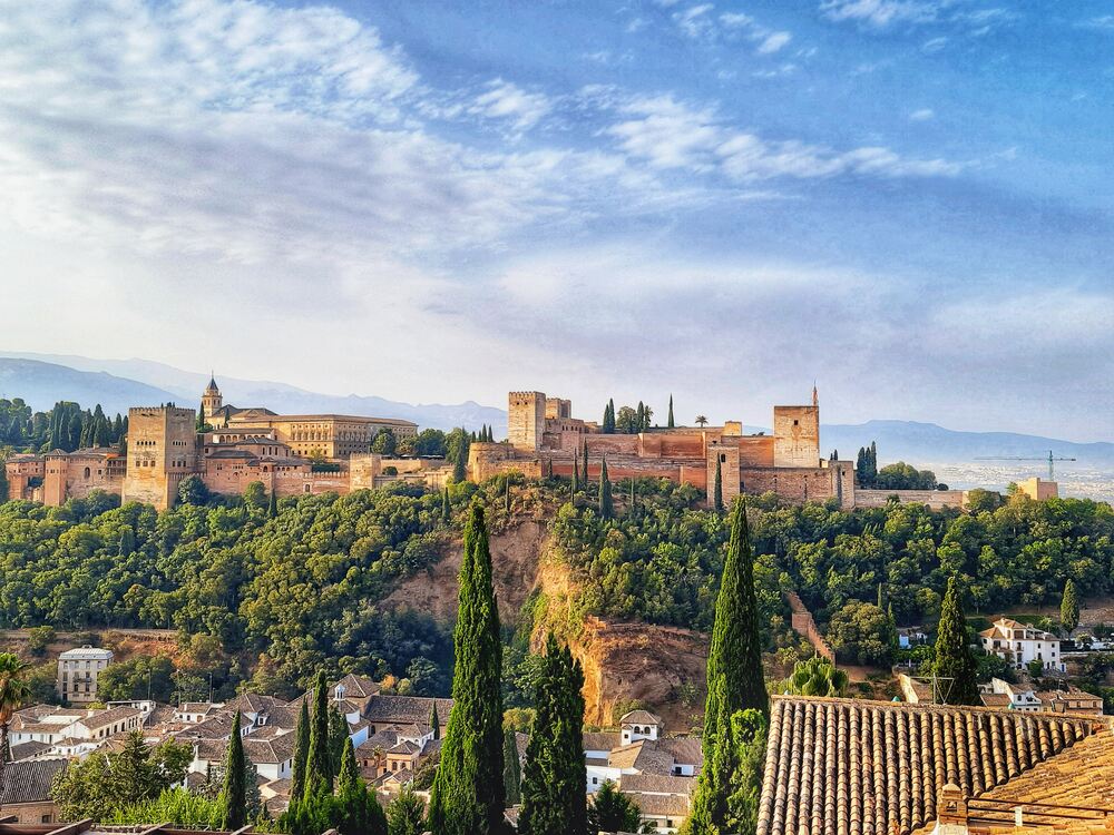 A view of the Alhambra in Granada, Spain. The fortress sits on a hill top, surrounded by trees and with houses in the bottom of the valley. You can see the Sierra Nevada mountains in teh background. I took this photo from Albaicin District at sunrise.