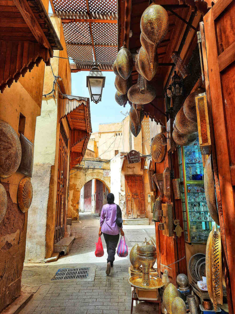 A photo of a woman wearing a purple top and black head scarf. She is carrying two bags of produce and walkting though the streets of Fes in Morocco.