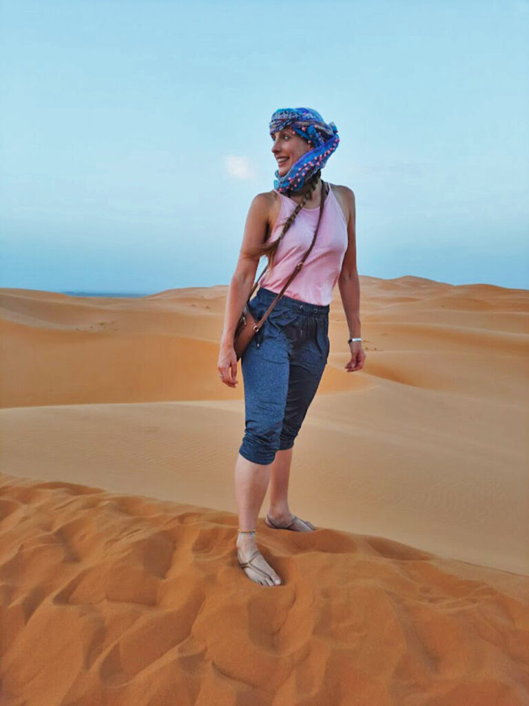 Me stood on top of a sand dune in the desert in Morocco. I am wearing a blue and pink turban and matching pink top. I'm looking out in to the distance over my right shoulder at dusk.