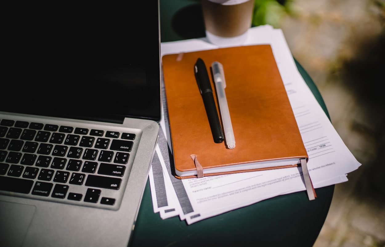 A brown notebook and pile of paper sitting on a table next to a silver laptop