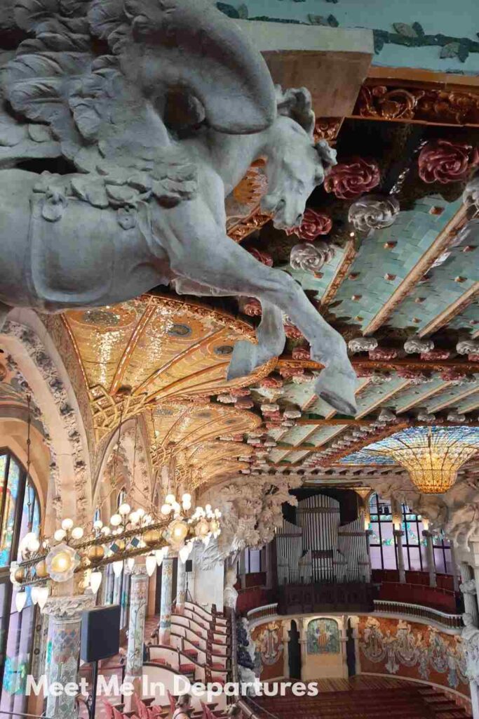 Palau de la Música Catalana Interior of the Palau de la Música Catalana in Barcelona, featuring ornate columns, colourful stained glass, sculpted figures including a dramatic horse relief, and a domed skylight ceiling.