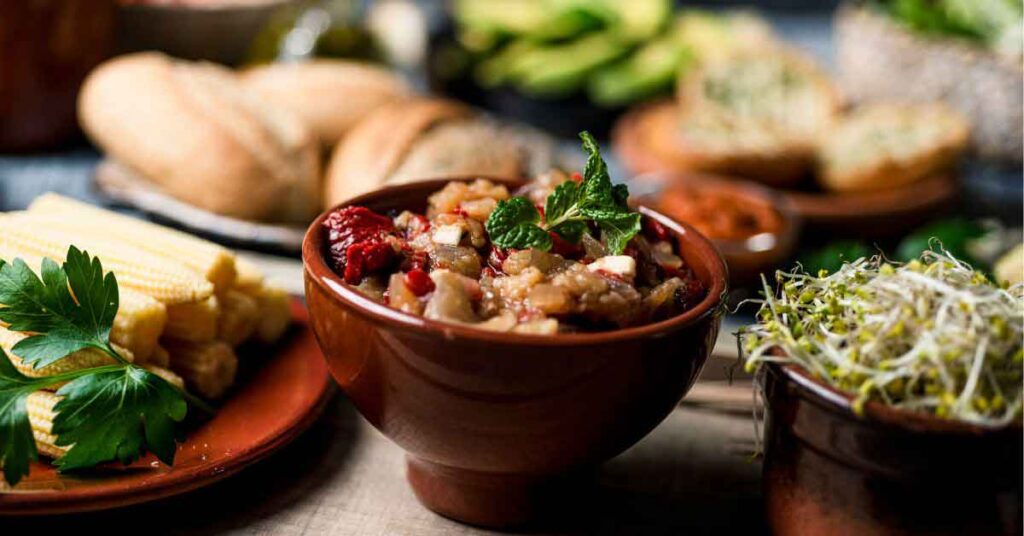 A rustic wooden table spread with various traditional dishes featuring vegetarian food in Spain. The centerpiece is a clay bowl filled with a hearty eggplant and vegetable stew garnished with fresh mint, surrounded by baby corn, sprouts, and crusty bread.