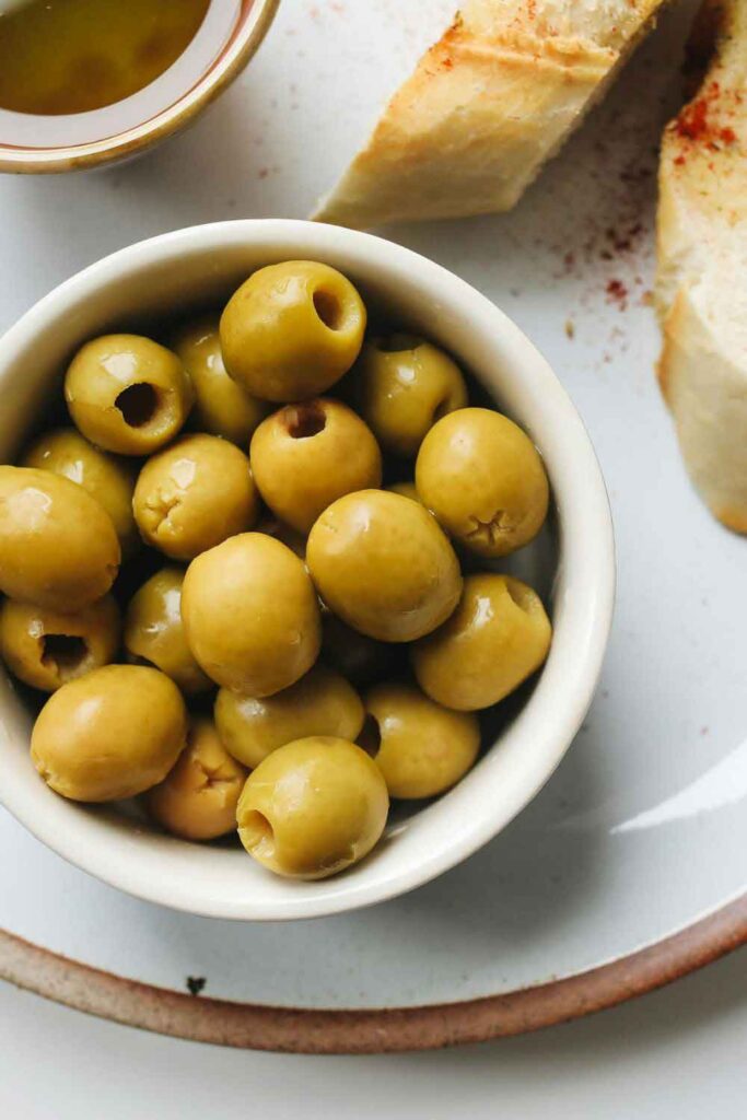 Bowl of green olives served alongside crusty bread and olive oil on a ceramic plate. A common start to a meat-free meal in Spanish dining.