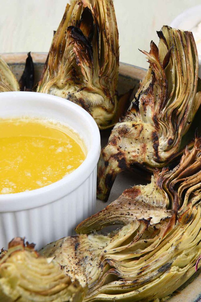 Charred artichoke halves surrounding a ramekin of melted butter. This seasonal vegetable dish is a celebrated example of plant-forward eating in Spain.