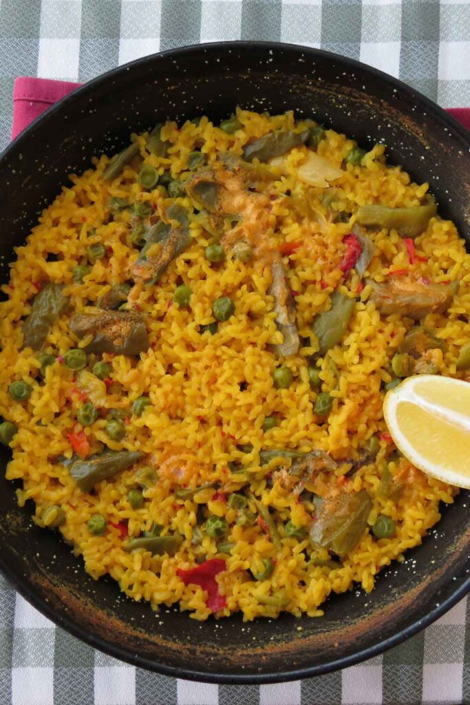 Overhead view of vegetable paella with saffron rice, artichokes, peas, and red peppers, garnished with a lemon wedge. A classic rice dish that showcases Spain’s vegetarian-friendly flavors.