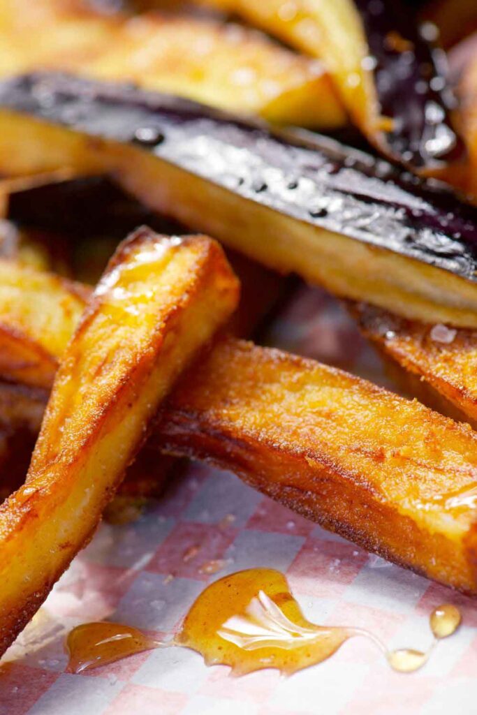 Crispy sticks of fried eggplant and potato drizzled with honey on checkered paper. A sweet and savory tapa that’s part of traditional meatless dining in Spain.