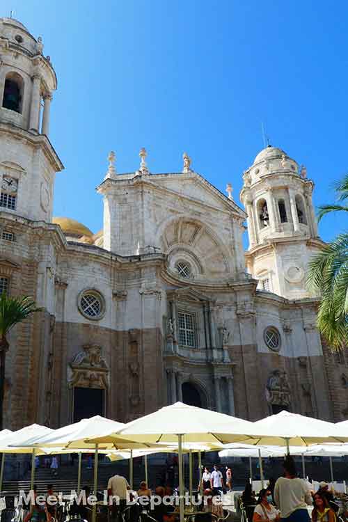The grand facade of Cádiz Cathedral in Spain stands tall with its twin towers and ornate baroque architecture. Below, people sit under white umbrellas in a sunny plaza lined with palm trees.
