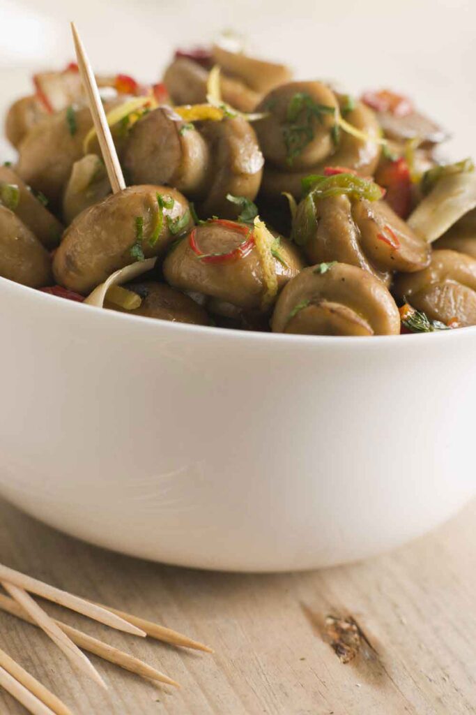 Bowl of marinated mushrooms with herbs, chili, and toothpicks for serving. A flavorful tapa often enjoyed in Spain’s meatless appetizer offerings.