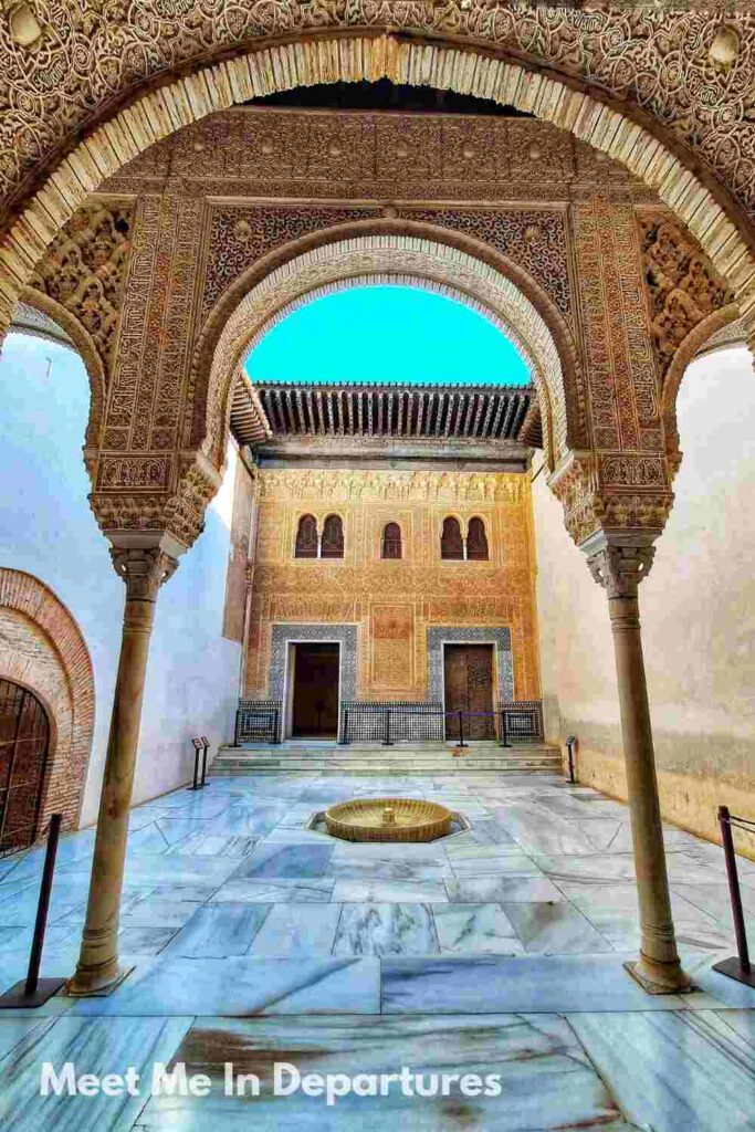 Nasrid Palace Patios, Granada Elegant courtyard in the Alhambra with carved archways, a central marble fountain, and golden stucco detailing on the building’s facade beneath a vivid blue sky.