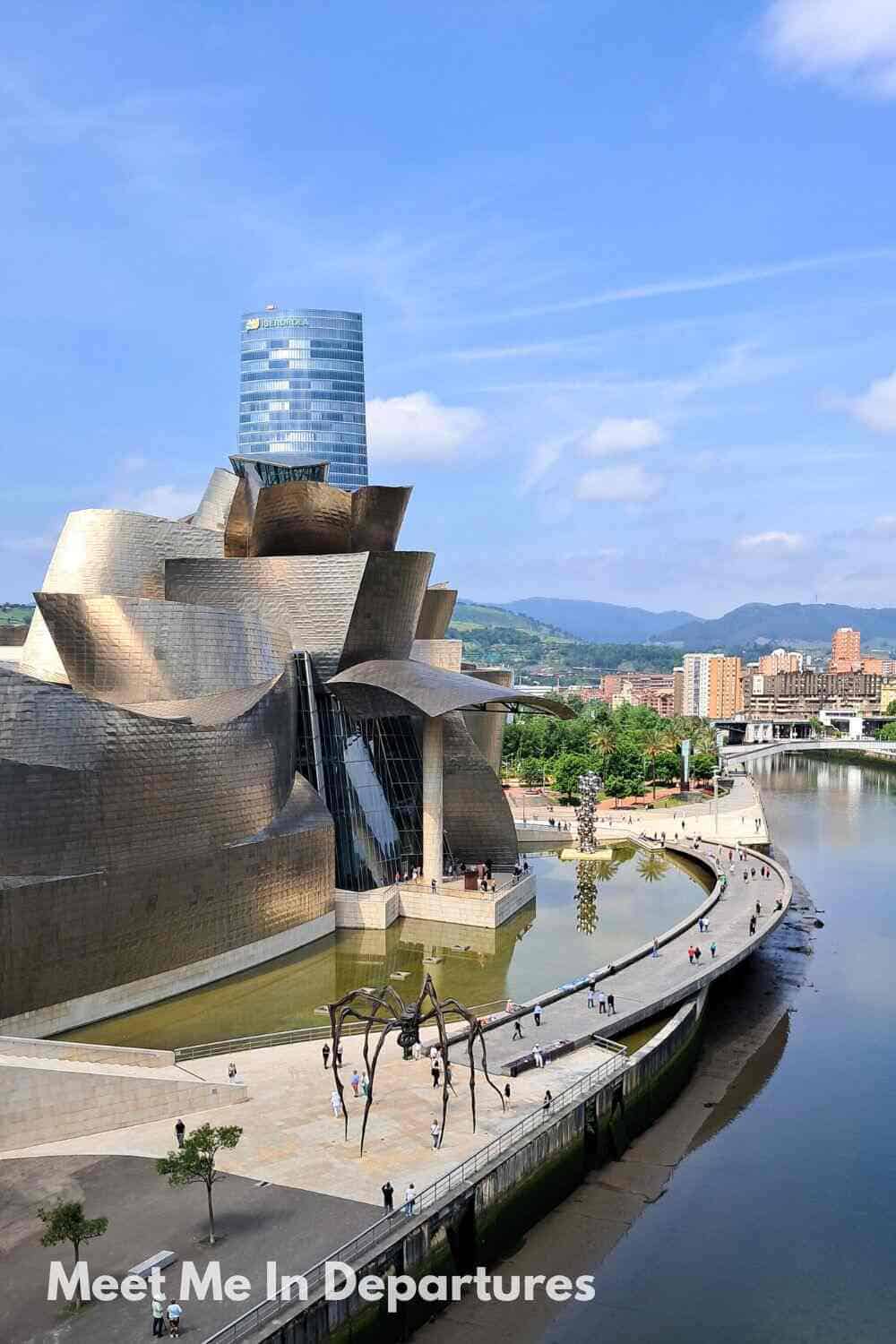 The iconic titanium-clad exterior of the Guggenheim Museum Bilbao reflecting sunlight beside the Nervión River, with Louise Bourgeois’s giant spider sculpture, Maman, standing on the walkway and a modern skyscraper rising in the background.