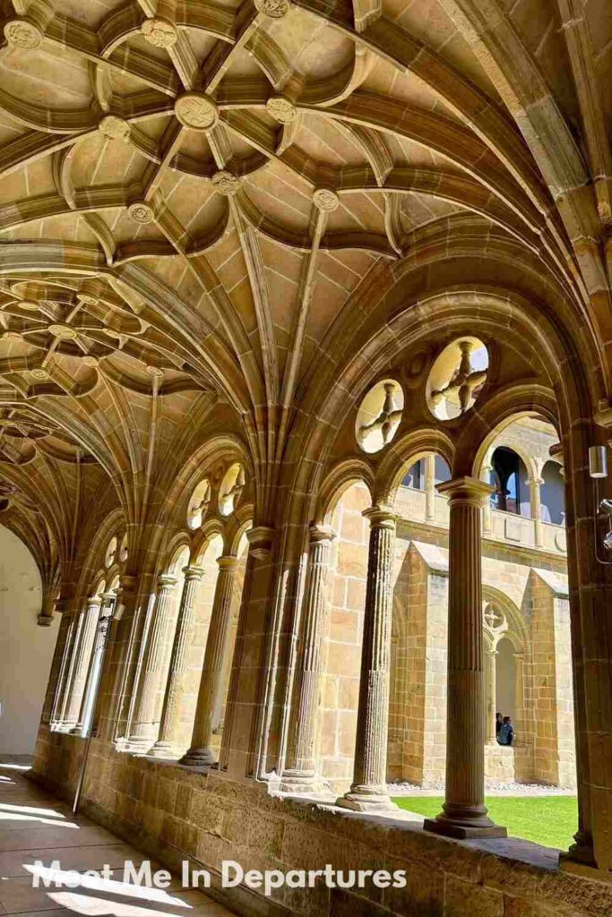 Arched stone cloister with ribbed vaulted ceilings and ornate columns, leading to a sunny courtyard in a historic monastery or cathedral.