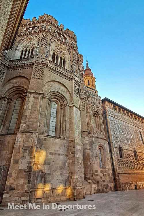 La Seo of Zaragoza Towering stone facade of a Mudejar-style cathedral in Aragón, Spain, with intricate brick and tile patterns lit by golden hour sunlight against a clear blue sky.