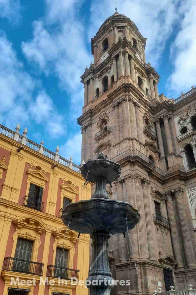 Ornate fountain in front of the Málaga Cathedral, with its towering bell tower and baroque stonework rising behind vibrant yellow and terracotta buildings under a partly cloudy sky.