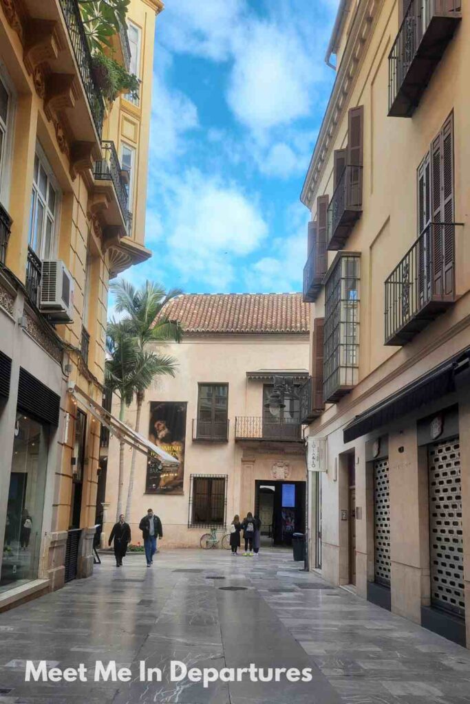 Quiet pedestrian street in Málaga lined with traditional buildings and balconies, leading toward the Carmen Thyssen entrance with a visible exhibition banner and a few people walking nearby.