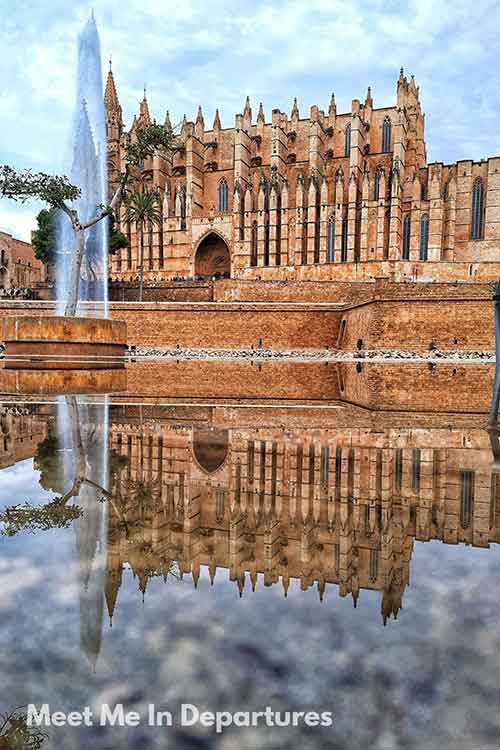 Palma de Mallorca Cathedral Reflection The sandstone facade of La Seu Cathedral in Palma de Mallorca, Spain, is dramatically reflected in a large pool of water, with a tall fountain spraying in the foreground. The Gothic architecture is framed by a cloudy sky and stone walls.