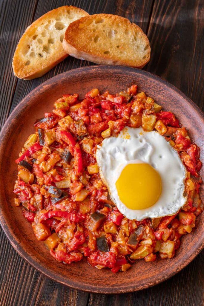 Pisto manchego, a mix of sautéed zucchini, tomato, and peppers, topped with a sunny-side-up egg and toasted bread. A hearty vegetable dish often enjoyed meat-free.