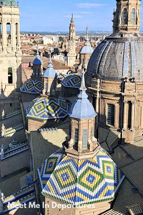 Rooftop of Basilica del Pilar Zaragoza Elevated view of the Basílica del Pilar in Zaragoza, highlighting its colorful, geometric-tiled domes surrounded by spires and the cityscape in the background.