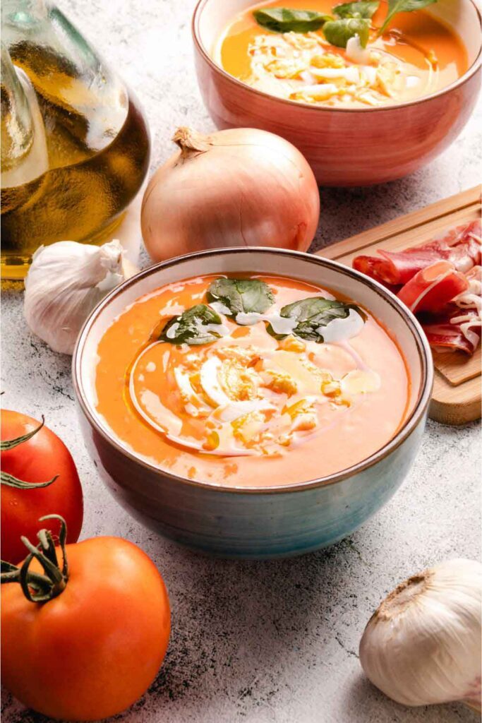 Bowl of creamy salmorejo garnished with chopped boiled egg and olive oil, surrounded by tomatoes and garlic. A classic cold soup that's a favorite in Spanish vegetarian cooking.