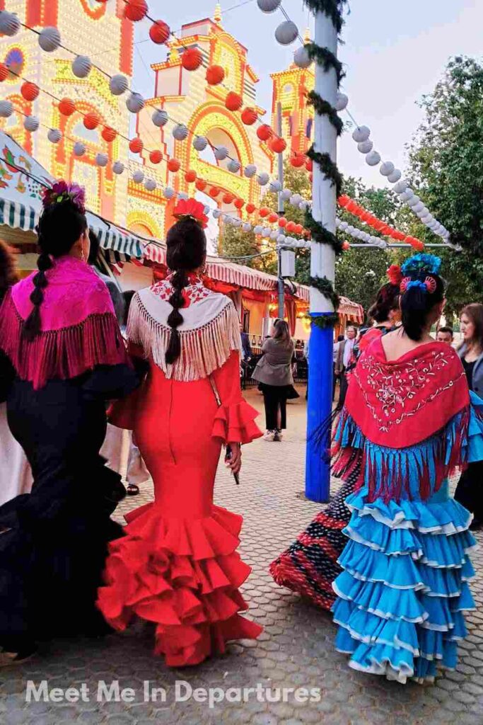 Women wearing vibrant flamenco dresses with ruffled skirts and shawls walk through Seville’s Feria de Abril, with colorful paper lanterns strung overhead and illuminated fairground arches in the background.