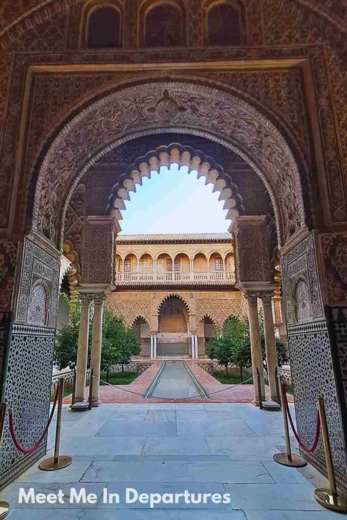 Ornate Moorish archway framing the view of a courtyard garden and reflecting pool inside the Real Alcázar of Seville, showcasing intricate Islamic architectural details and tilework.