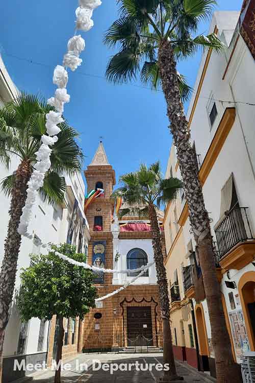 A narrow cobblestone street in Cádiz, Spain, lined with tall palm trees and white streamers overhead, leads to a small brick church with a bell tower and colorful flags. The church is framed by white and yellow buildings under a clear blue sky.