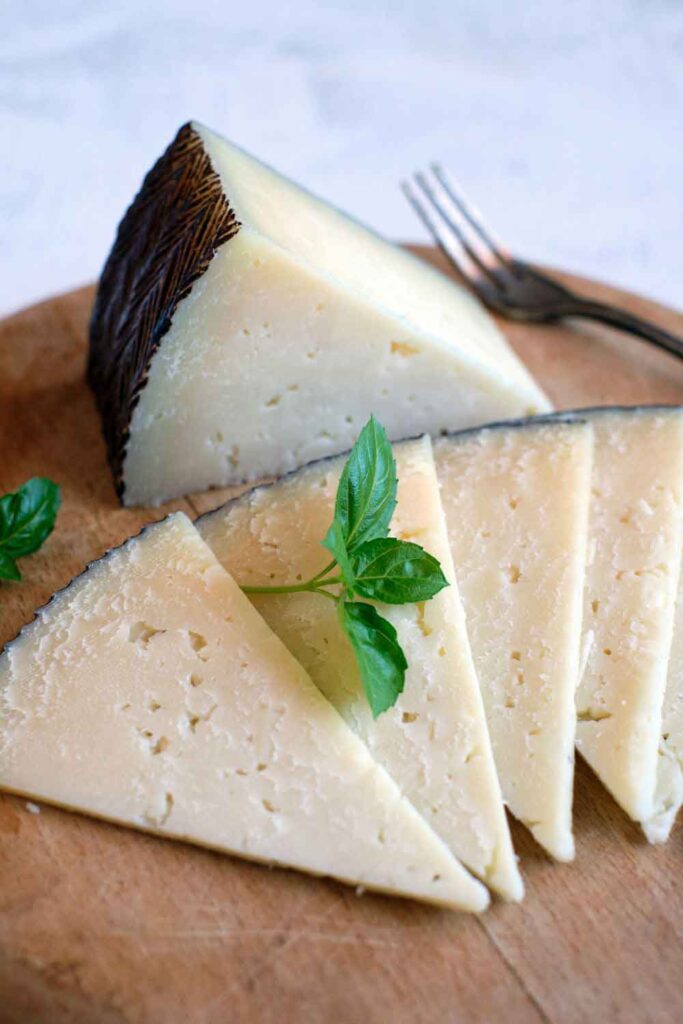 Wedge and slices of aged manchego cheese garnished with a basil sprig. A key ingredient in many meat-free Spanish tapas platters.