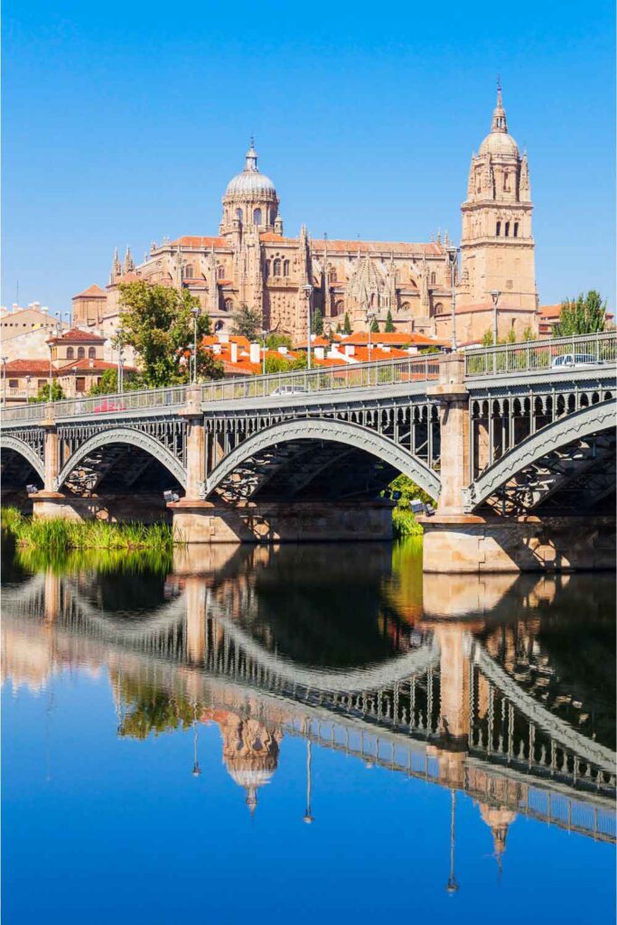 Salamanca Cathedral View of Salamanca Cathedral with its grand towers and dome, reflected in the calm waters of the Tormes River beneath a historic iron bridge.