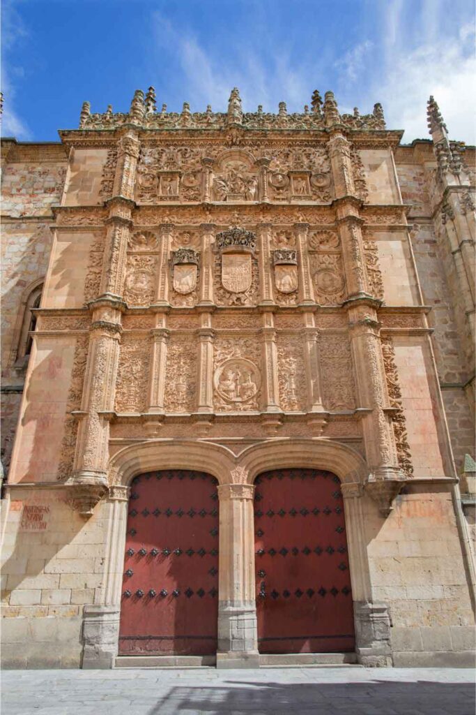 Salamanca University Intricately carved Plateresque façade of the University of Salamanca, featuring coats of arms, decorative stonework, and twin red doors under a blue sky.