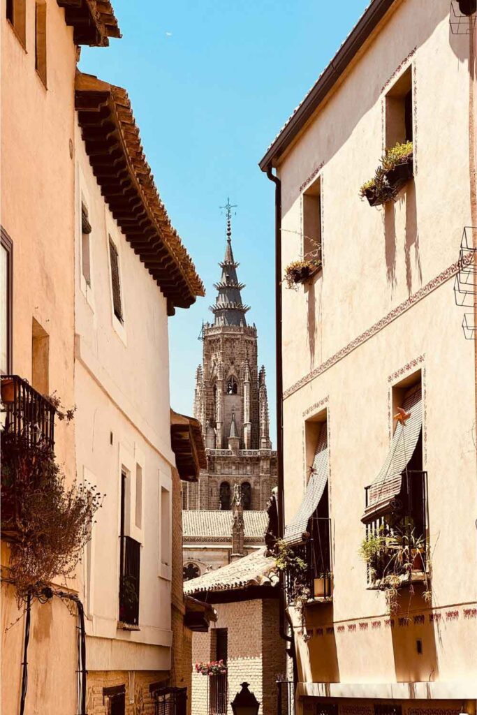 Toledo Cathedral Narrow cobbled street in Toledo framed by traditional buildings with flower boxes, looking towards the towering spire of Toledo Cathedral in the background.