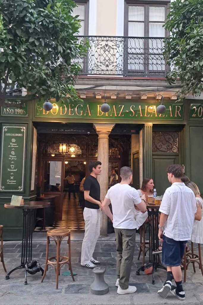 Tapas Bars in Seville Street view of Bodega Díaz-Salazar in Seville, with its green facade, gold lettering, and classic tiled entrance dating back to 1908. A small group of people stand around high tables outside, enjoying drinks in the lively tapas bar atmosphere.
