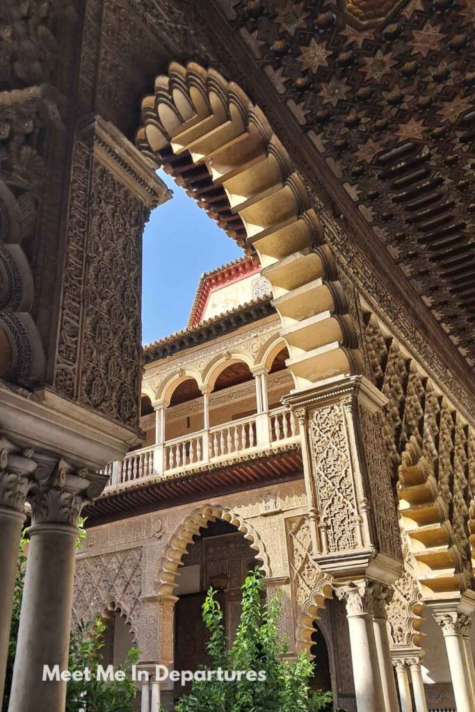 View through an ornate horseshoe arch towards the upper galleries of the Patio de las Doncellas at the Real Alcázar of Seville, highlighting intricate Mudejar stucco work and lush courtyard greenery, taken during a Alone in the Alcazar VIP tour.