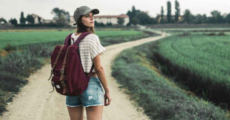 A young woman wearing a striped crop top, denim shorts, and a red backpack walks alone along a winding dirt path through green fields in rural Spain. She looks over her shoulder, appearing calm and confident. This peaceful countryside scene reflects the question: "Is Spain Safe for Solo Female Travellers?" while visually suggesting independence and thoughtful exploration.