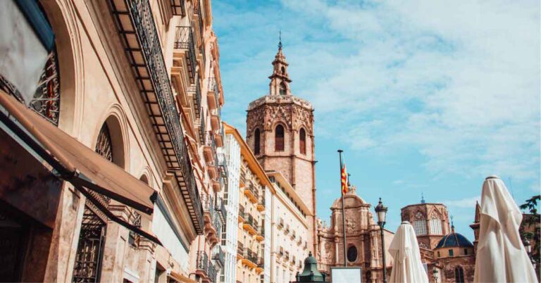 View of historic rooftops and architecture in Valencia, Spain, with the iconic Micalet bell tower rising above sunlit buildings and café umbrellas in the foreground. A charming cityscape that invites travelers to ask, is Spain worth visiting.