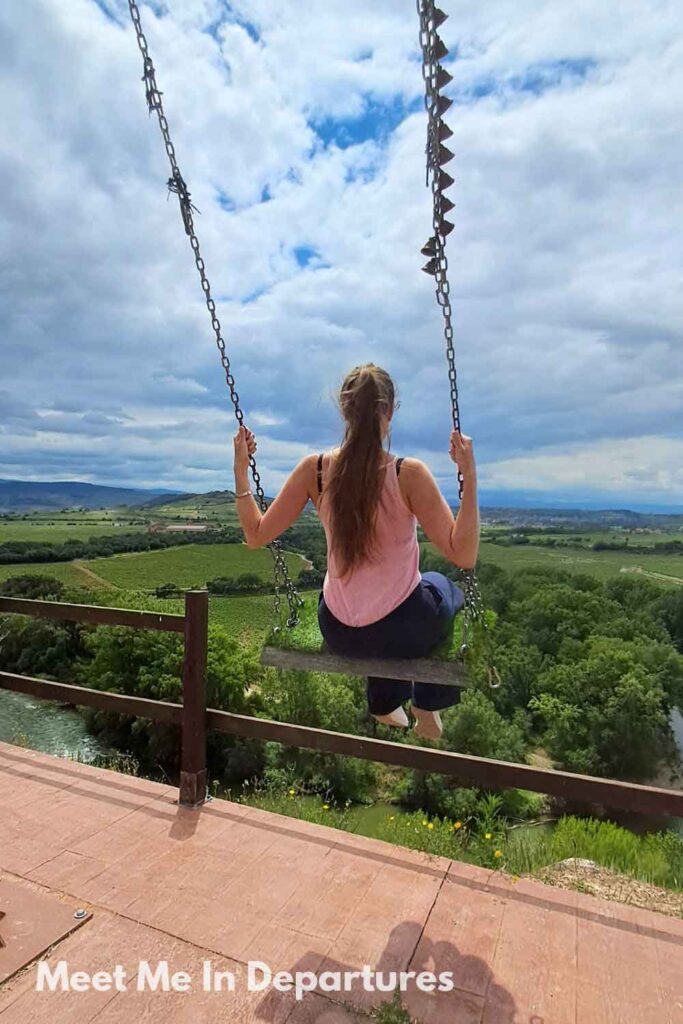 Woman (me) sitting on a swing overlooking a vast green valley, the Ebro River and vineyard landscape in the La Rioja region under a partly cloudy sky in northern Spain. A peaceful moment capturing the beauty and freedom of rural Spanish countryside.