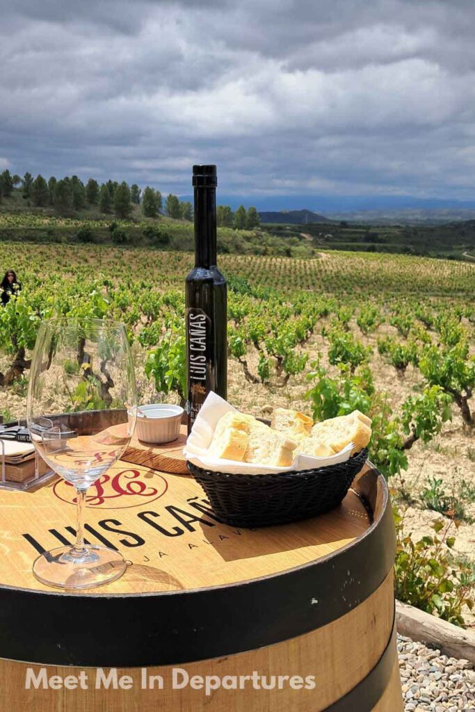 Vineyards in La Rioja Region Basket of bread, an empty wine glass, and a bottle of Luis Cañas wine resting on a barrel table in the middle of a sunlit vineyard in Rioja Alavesa. A scenic setup for wine tasting surrounded by rolling green vines and dramatic skies in northern Spain.