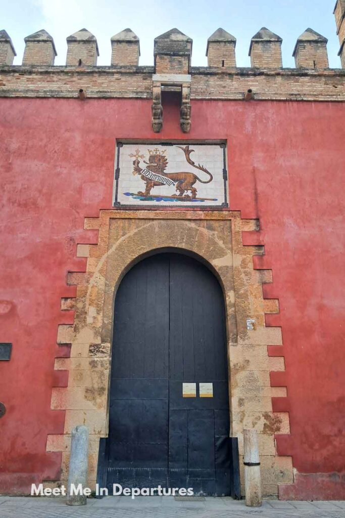 Close-up view of the Puerta del León (Real Alcázar de Sevilla), showing the arched stone doorway set in a red wall with a tile of a crowned lion holding a cross above the entrance, leading inside the Real Alcázar of Seville.