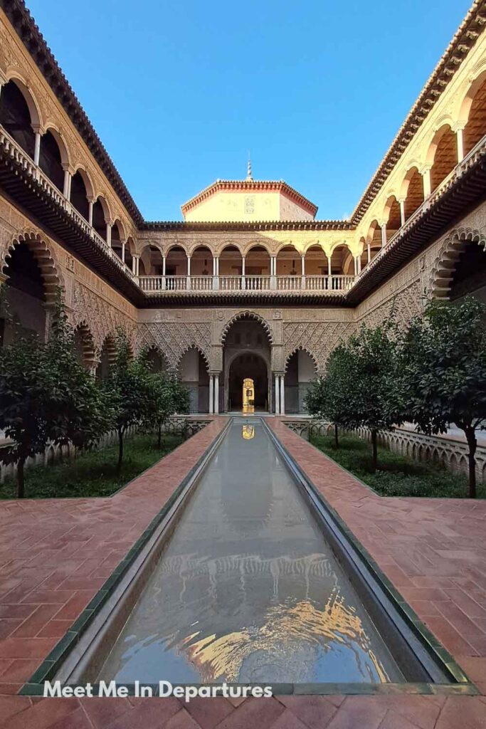 Symmetrical view of the Patio de las Doncellas at the Real Alcázar of Seville, with a long reflective pool, orange trees, and two-storey arched galleries, taken during early morning access before visitors arrived.