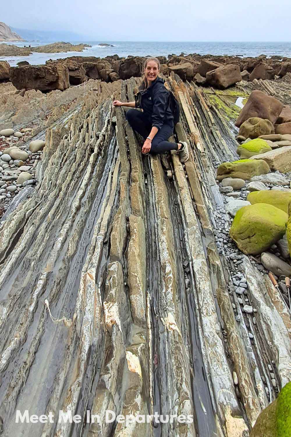 Woman (me) crouching on dramatic layered flysch rock formations along the Basque Coast, with the ocean and rocky shoreline in the background. This unique geological landscape is part of northern Spain’s striking natural scenery.