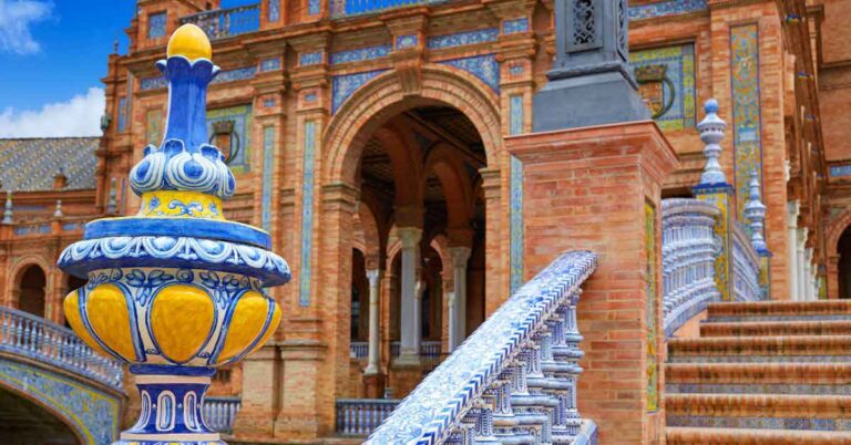 Close-up of colourful blue, yellow, and white ceramic tiles on a bridge at Plaza de España in Seville, with the ornate brick archways and tiled columns of the building in the background – a perfect example of why Seville is one of the best cities in southern Spain to visit.