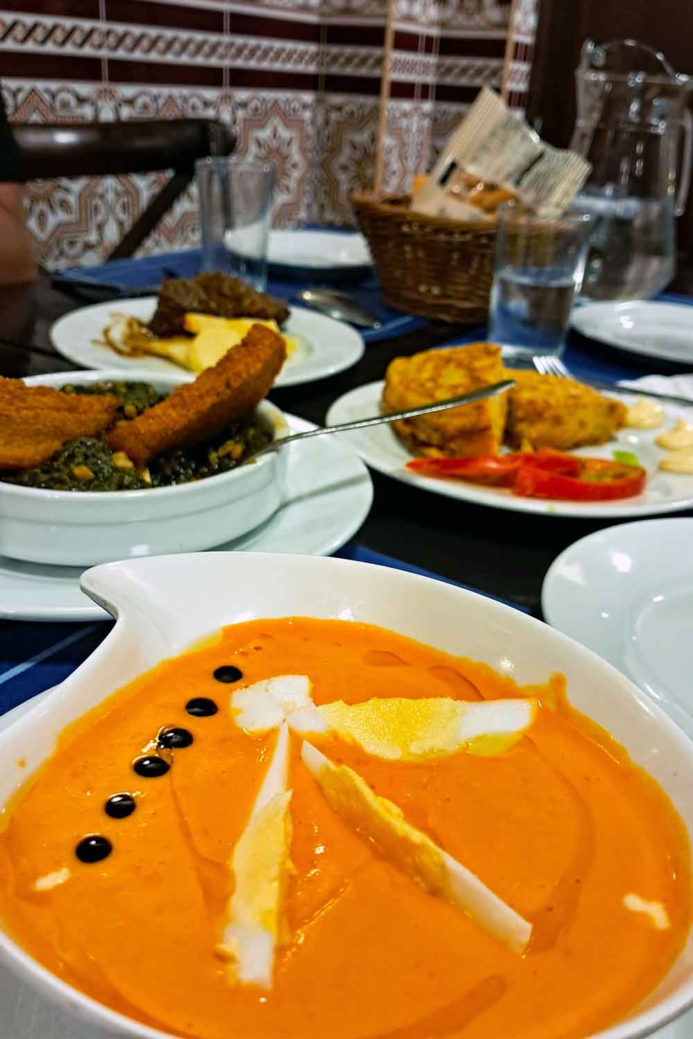 Close-up of a creamy bowl of salmorejo topped with slices of hard-boiled egg and balsamic drops, served alongside plates of Spanish tortilla, fried fish, and other tapas on a table in Seville. The background features traditional Andalusian tilework, adding to the authentic dining atmosphere.