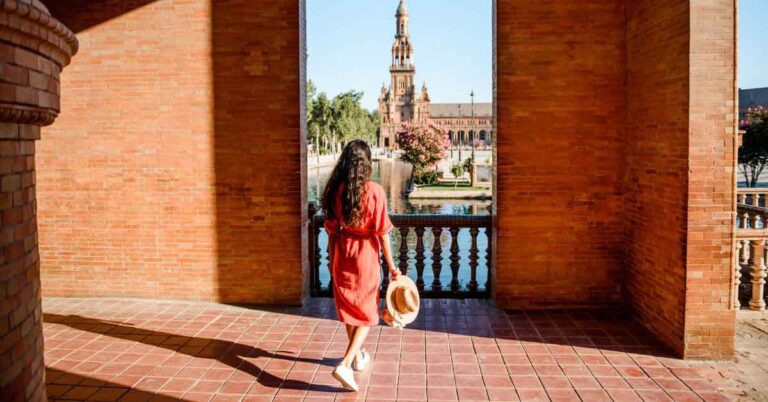 Alt text: A woman in a red dress holding a hat walks towards the scenic Plaza de España in Seville, Spain, framed by red brick arches and a view of the canal and tower — a perfect inspiration for anyone researching how to plan a trip to Spain.