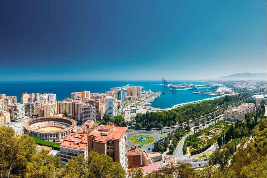 Malaga from above A panoramic view of Malaga, Spain, showing the city's bullring, modern buildings, lush green park, and the port with the deep blue Mediterranean Sea in the background.