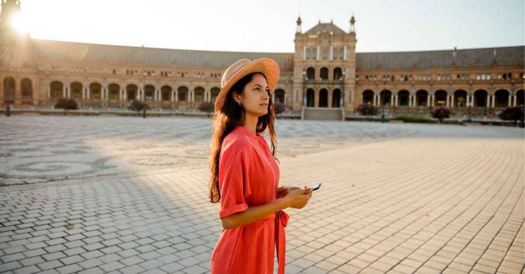 A woman in a coral dress and straw hat stands in the sunlit Plaza de España in Seville, holding her phone and taking in the view. The image reflects ideas from a Spain packing list and shows what to wear in Spain for warm, sunny days.