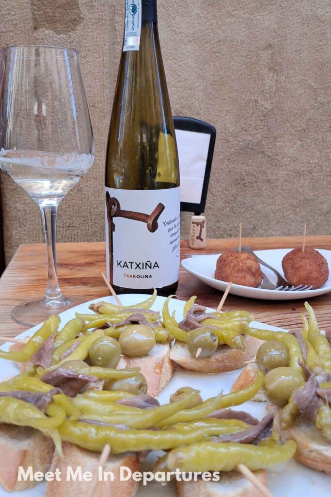 Plate of pintxos with green peppers, olives, and anchovies, alongside a bottle of Txakoli wine at a bar in San Sebastian, showcasing the city’s renowned Basque cuisine.