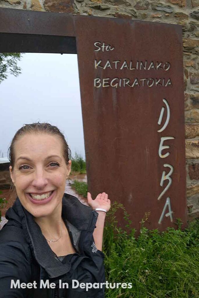 Smiling woman (me) in a rain jacket taking a selfie in front of the rust-colored entrance sign for Sta. Katalinako Begiratokia in Deba, Basque Country. A misty but scenic stop along a coastal hiking route in northern Spain.