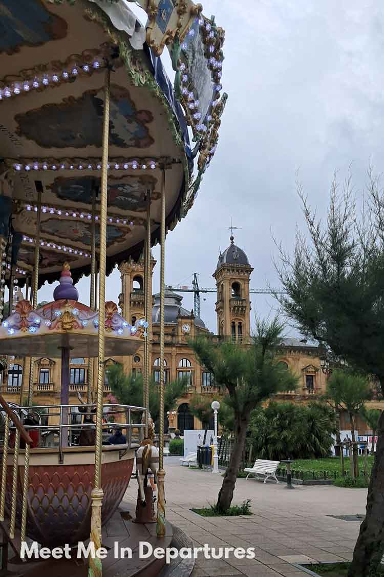 Colorful vintage carousel with pastel horses in the foreground and San Sebastián City Hall in the background under a cloudy sky. A playful and elegant scene in one of northern Spain’s most beloved cities.
