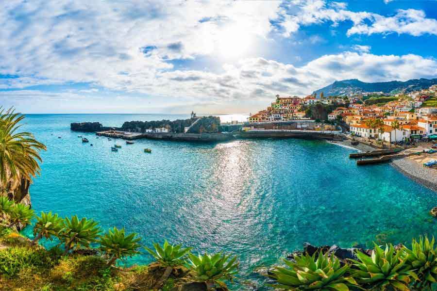 Câmara de Lobos, Madeira A stunning coastal view of Câmara de Lobos, Madeira, with turquoise water, colourful fishing boats, and whitewashed buildings with terracotta roofs nestled on the hillside. Lush greenery in the foreground and the sun reflecting off the sea highlight why this is one of the most scenic places to enjoy mild winter weather in southern Europe.