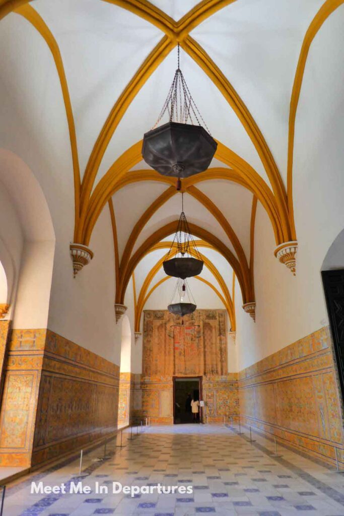 Interior view of the Chapel of the Gothic Palace inside the Real Alcázar of Seville, featuring high vaulted ceilings with golden ribbed arches, hanging lanterns, and walls decorated with colourful tiles leading to a doorway with a large tapestry above it.